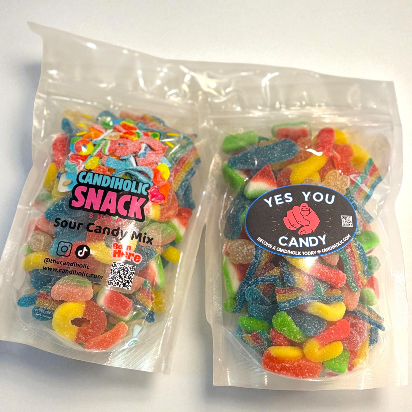 Two clear resealable bags filled with colorful sour gummy candies, including rainbow belts, gummy rings, watermelon slices, and other assorted shapes. The left bag has a bright “Candiholic Snack” label with social media icons, while the right bag has a round “Yes You Candy” label with a pointing hand graphic