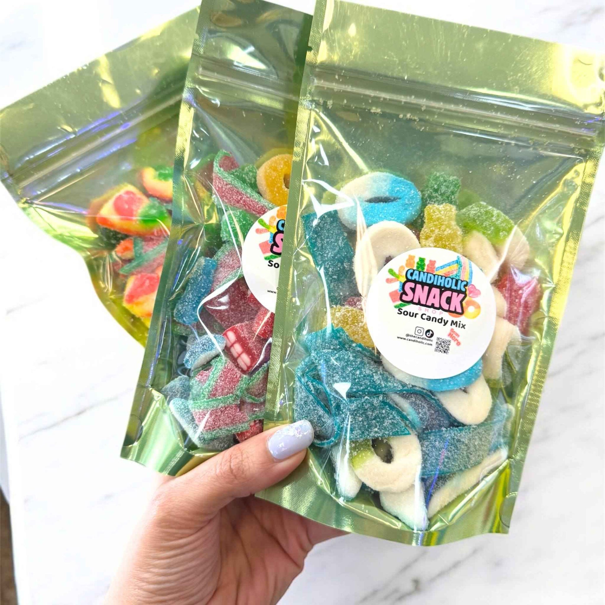 Bag of sour candy mix with rainbow belts, gummy rings, and worms under bright pink flowers